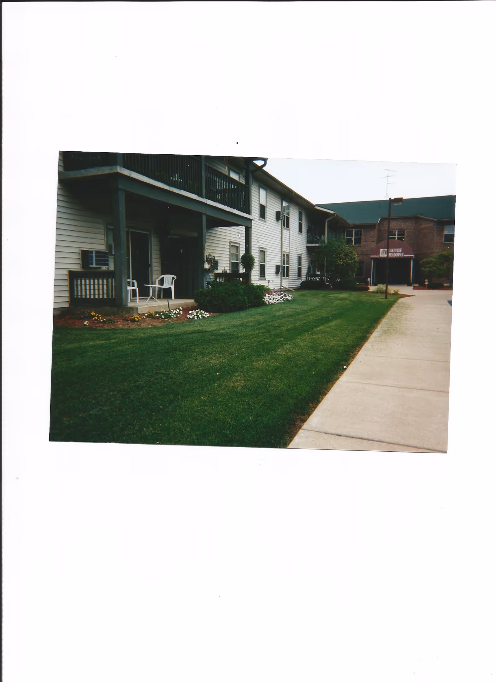 Exterior view of a senior living facility named Lakeview Meadows I, showing a two-story building with white siding and a brick entrance. There is a well-maintained lawn with flower beds and a concrete walkway leading to the entrance. Two white chairs and a small table are placed on a covered porch area.