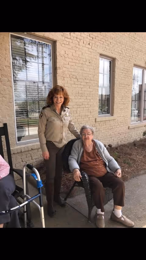 An elderly woman sitting on a wicker chair outside a brick building, accompanied by a smiling woman in a uniform standing beside her. A walker is visible next to them.