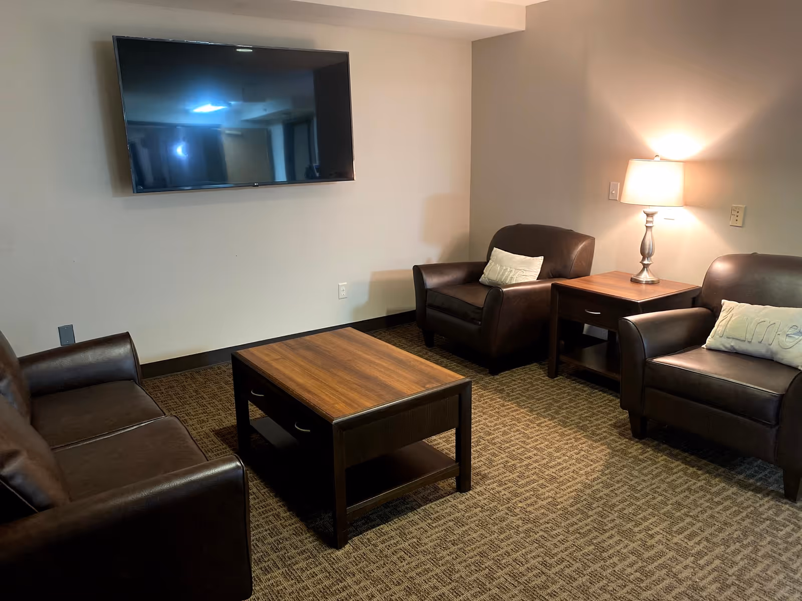 A cozy living room area with a wall-mounted flat-screen TV, a brown leather sofa, two matching leather armchairs each with a light-colored pillow, a wooden coffee table in the center, and a wooden side table with a lit table lamp.