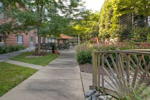 A paved walkway in an outdoor garden area of a senior living facility with green trees, flowering plants, a small wooden bridge, and outdoor seating with tables and chairs. The building is visible on the left side.