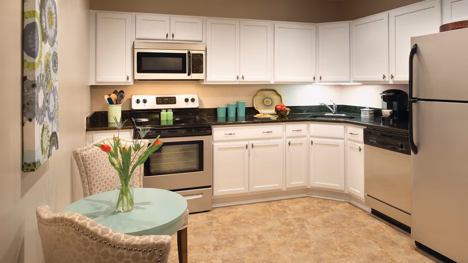 Small modern kitchen with white cabinets, stainless-steel appliances, black countertops, and a round table with a vase of tulips.
