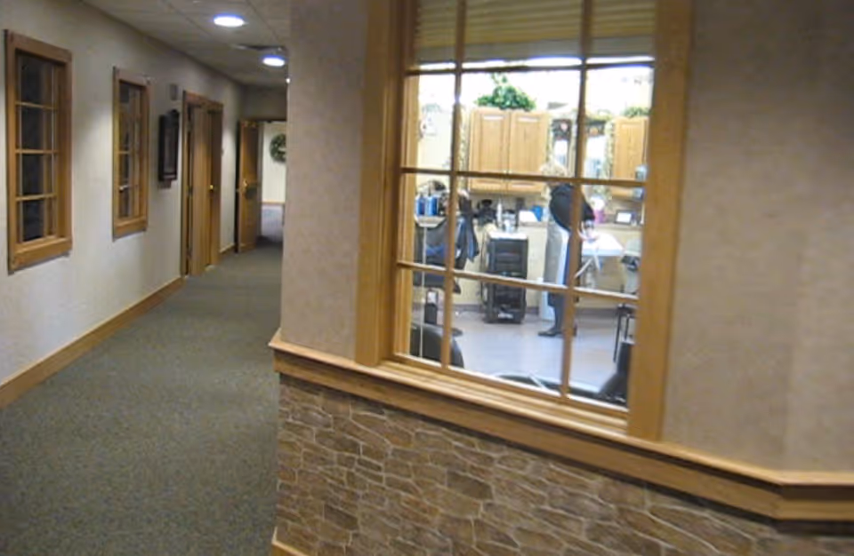 Carpeted interior hallway with doors and windows along the left and a large windowed partition revealing a staffed communal room.