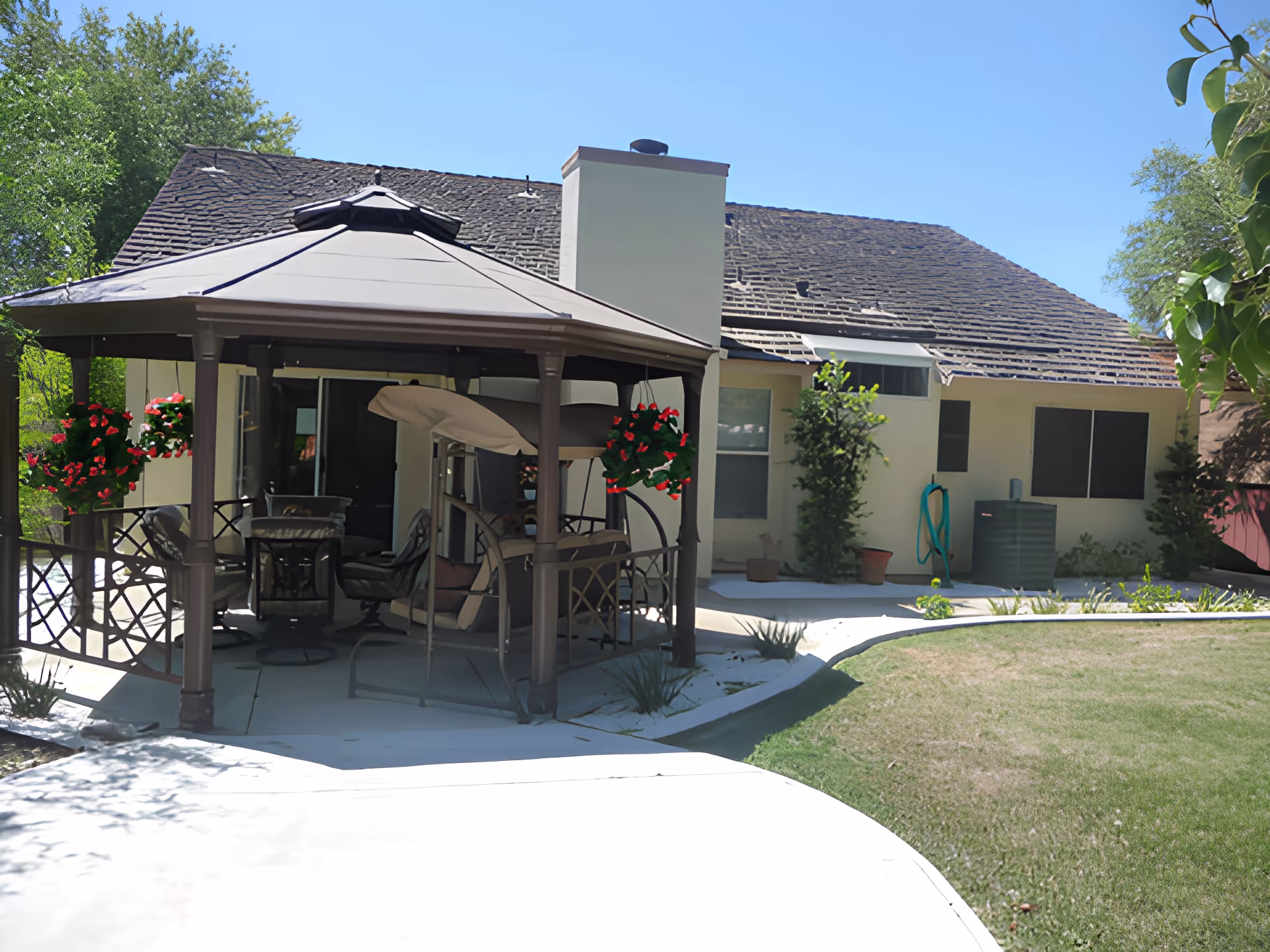 Outdoor patio area with a gazebo featuring cushioned chairs and a swing, surrounded by hanging flower baskets with red flowers. The patio is adjacent to a single-story building with a shingled roof, windows, and some greenery including small trees and potted plants. The sky is clear and blue.