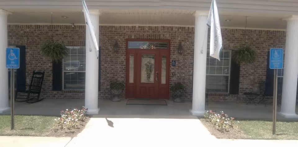 Front entrance of a brick building with a red door, two white columns, two handicap parking signs, hanging plants, and two flower beds on either side of the walkway.