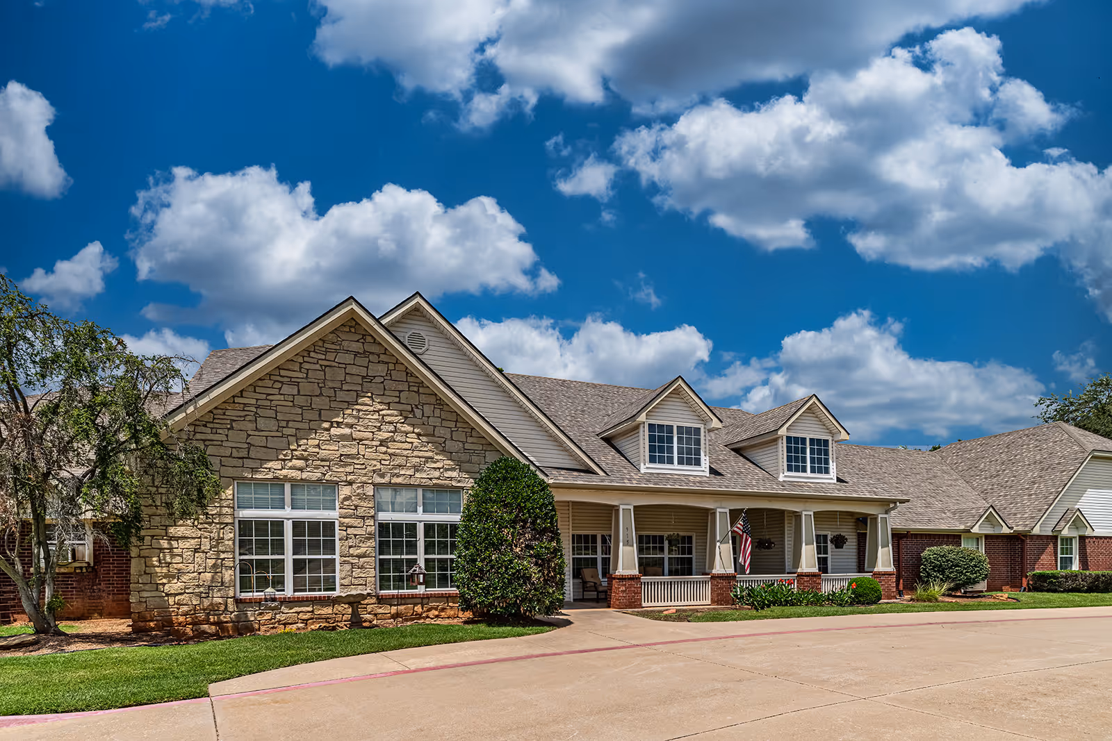 Exterior view of a single-story senior living facility building with stone and brick facade, multiple windows, a covered porch with columns, an American flag, and a well-maintained lawn under a blue sky with scattered clouds.