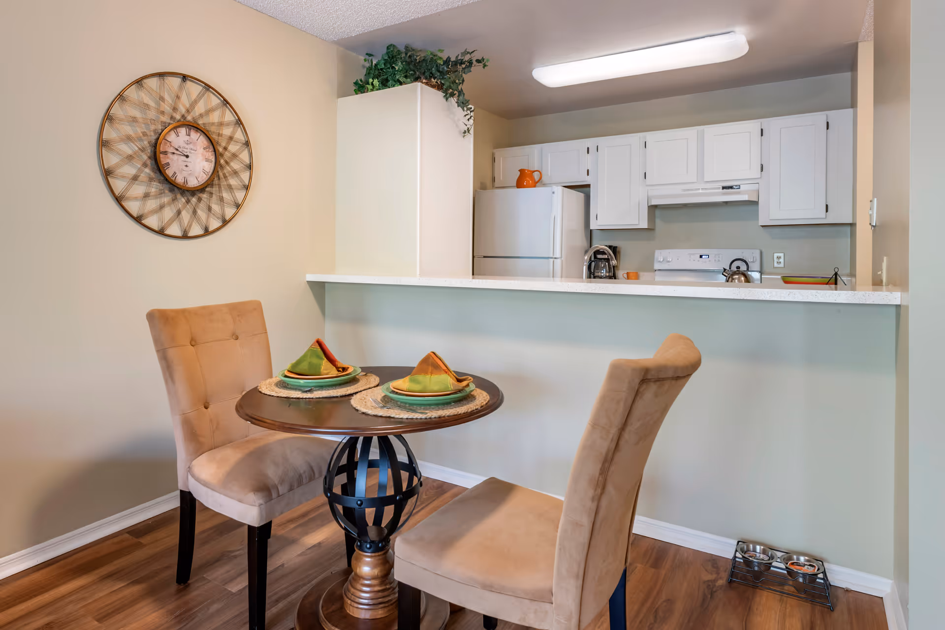 A small dining area with a round wooden table set for two with green and orange folded napkins on plates. Two beige upholstered chairs are positioned around the table. Behind the table is a kitchen with white cabinets, a white refrigerator, a stove, and a coffee maker. A decorative wall clock hangs on the beige wall to the left, and a small plant is placed on top of a cabinet. On the floor near the wall, there is a pet feeding station with two bowls.