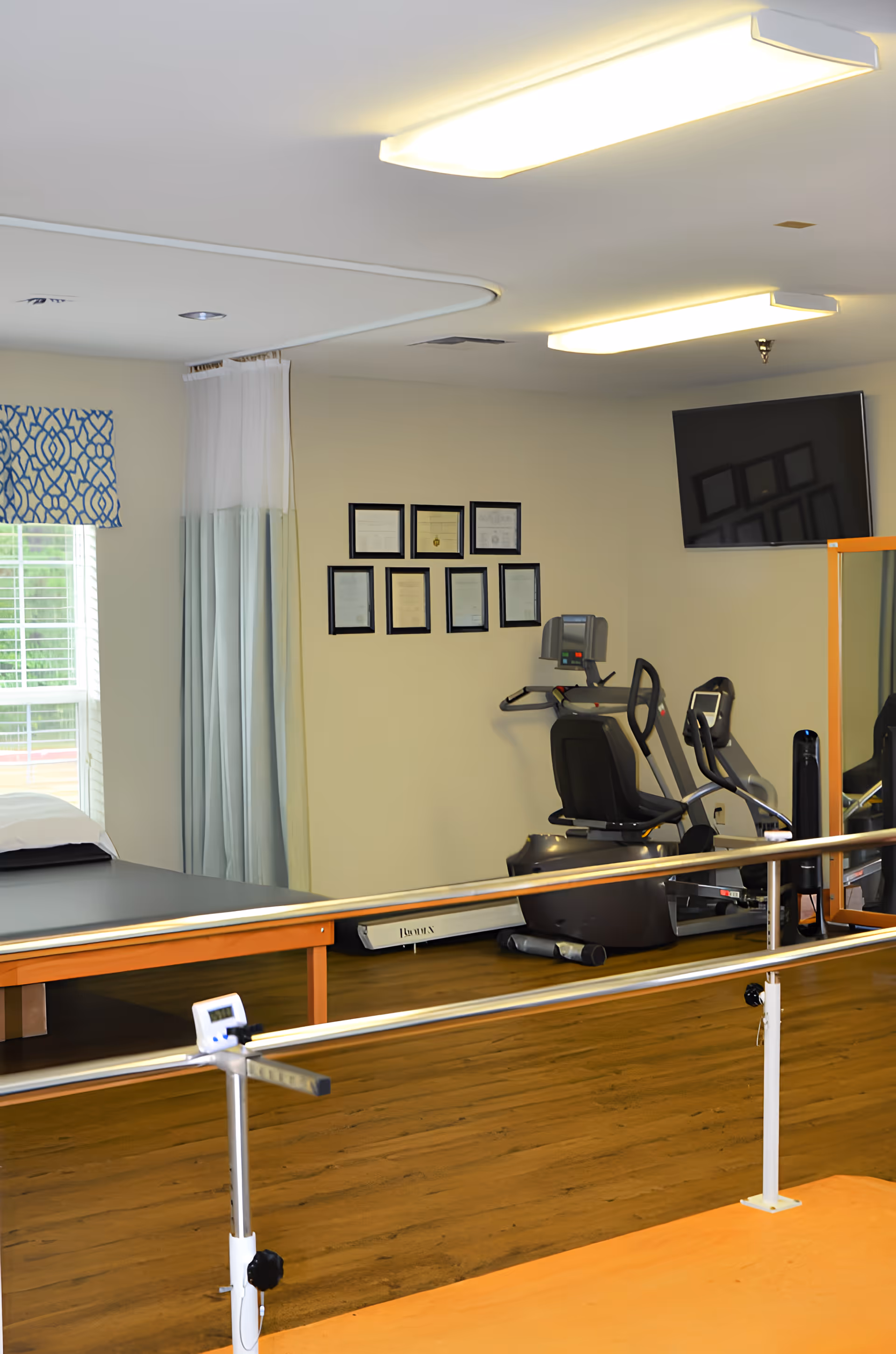 Interior therapy and fitness room with parallel walking bars, exercise equipment and a treatment table beneath framed certificates and a wall-mounted TV.