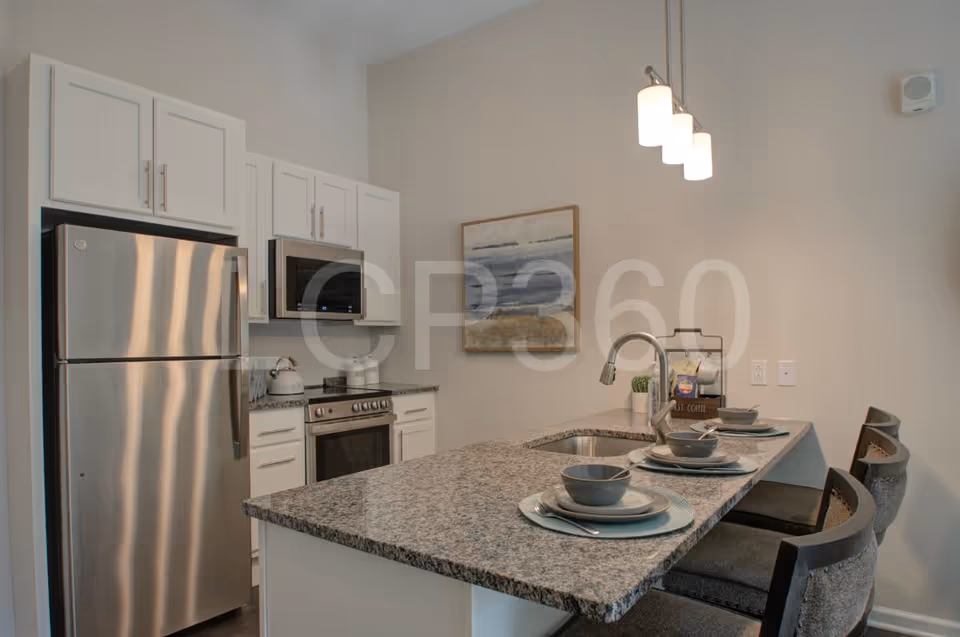 Modern kitchen with white cabinets, stainless steel refrigerator, oven, and microwave. A granite countertop island with a sink and three place settings with plates, bowls, and utensils. Three pendant lights hang above the island, and a framed abstract painting is on the wall.