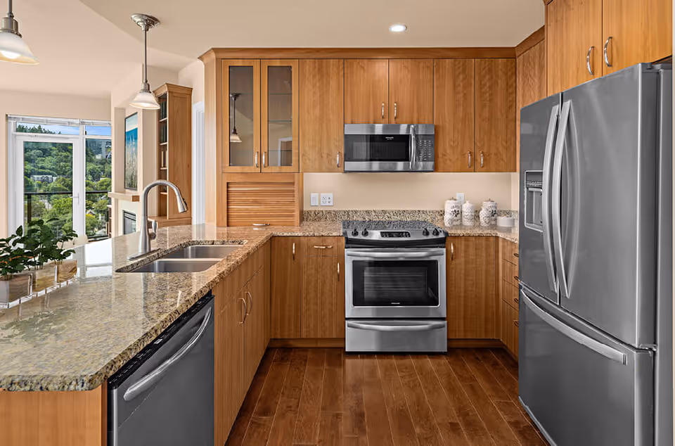 Bright modern kitchen with wooden cabinetry, granite countertops, stainless steel appliances, and a sink overlooking the living area.