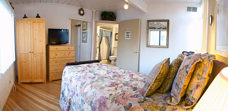A cozy bedroom with a floral-patterned bedspread and multiple pillows on the bed. The room features wooden flooring, a wooden wardrobe, and a chest of drawers with a flat-screen TV on top. A door is open, revealing a bathroom with a toilet and shower curtain. The walls are decorated with framed pictures and a mirror.