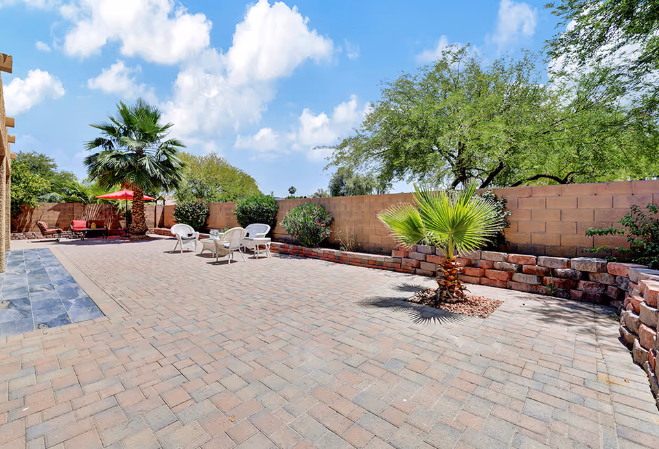 Outdoor patio area with brick pavers, several white chairs and a small table, surrounded by a low brick wall and various green plants including palm trees under a partly cloudy blue sky.