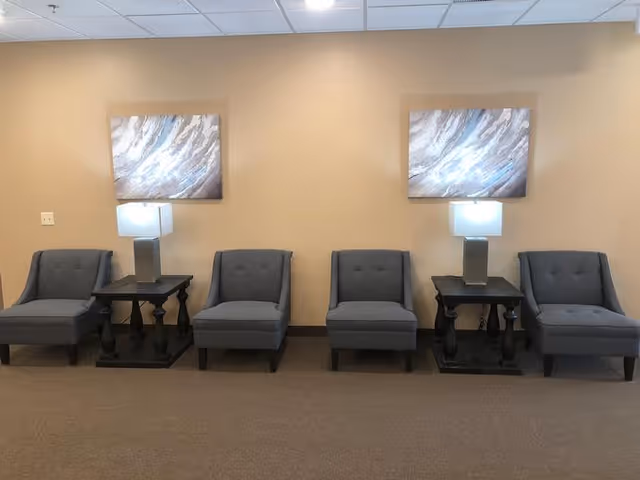 A row of four gray upholstered chairs separated by two black side tables with modern square lamps on top, set against a beige wall with two abstract paintings hanging above the lamps.
