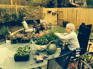 Two elderly women enjoying gardening together in a fenced backyard patio area. One woman is seated in a wheelchair while the other is sitting on a chair. They are surrounded by various plants, flowers, and gardening tools on a glass table.