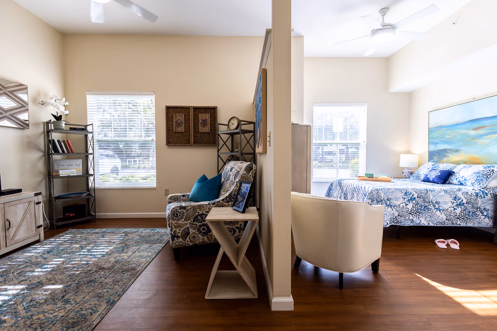 A bright and cozy living space divided by a partial wall. On the left side, there is a living room area with a patterned armchair, a small side table, a bookshelf with books and decor, a window with blinds, and a TV stand. On the right side, there is a bedroom area with a bed covered in blue and white patterned bedding, a white armchair, a nightstand with a lamp, a large painting above the bed, and a window with blinds. The floor is wooden throughout.