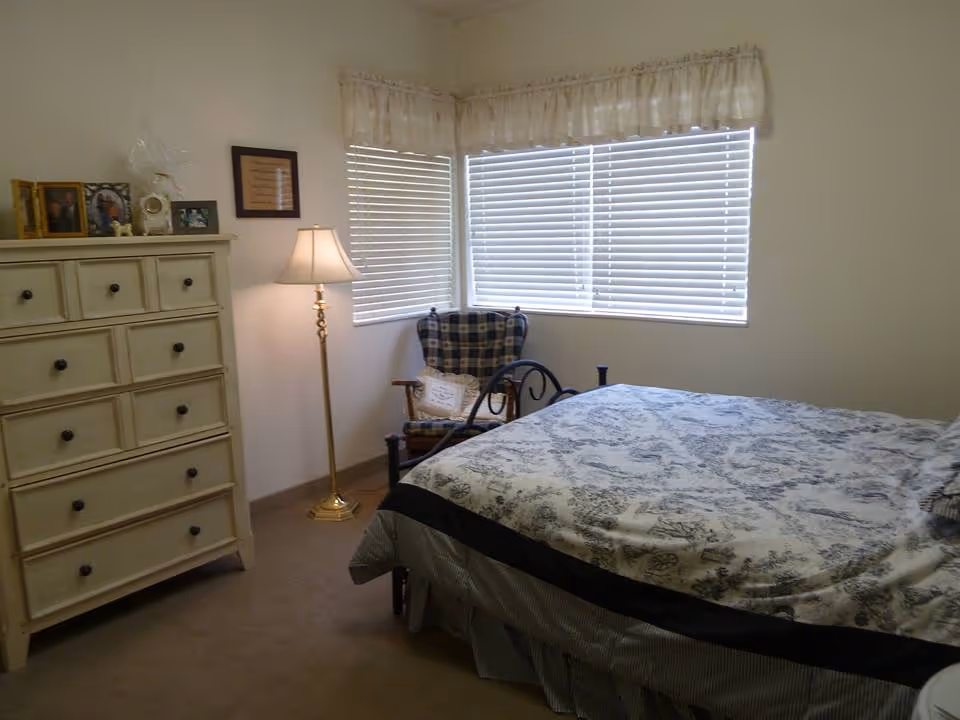 Bedroom with a patterned bed, a tall dresser, a floor lamp, and a plaid armchair by corner windows with blinds.