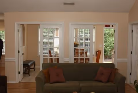 Interior view of a living room with a green sofa and two brown pillows. The room has light-colored walls and wooden flooring. Through two open doorways, a dining area with a wooden table and chairs is visible, along with windows that have white blinds and a view of greenery outside.