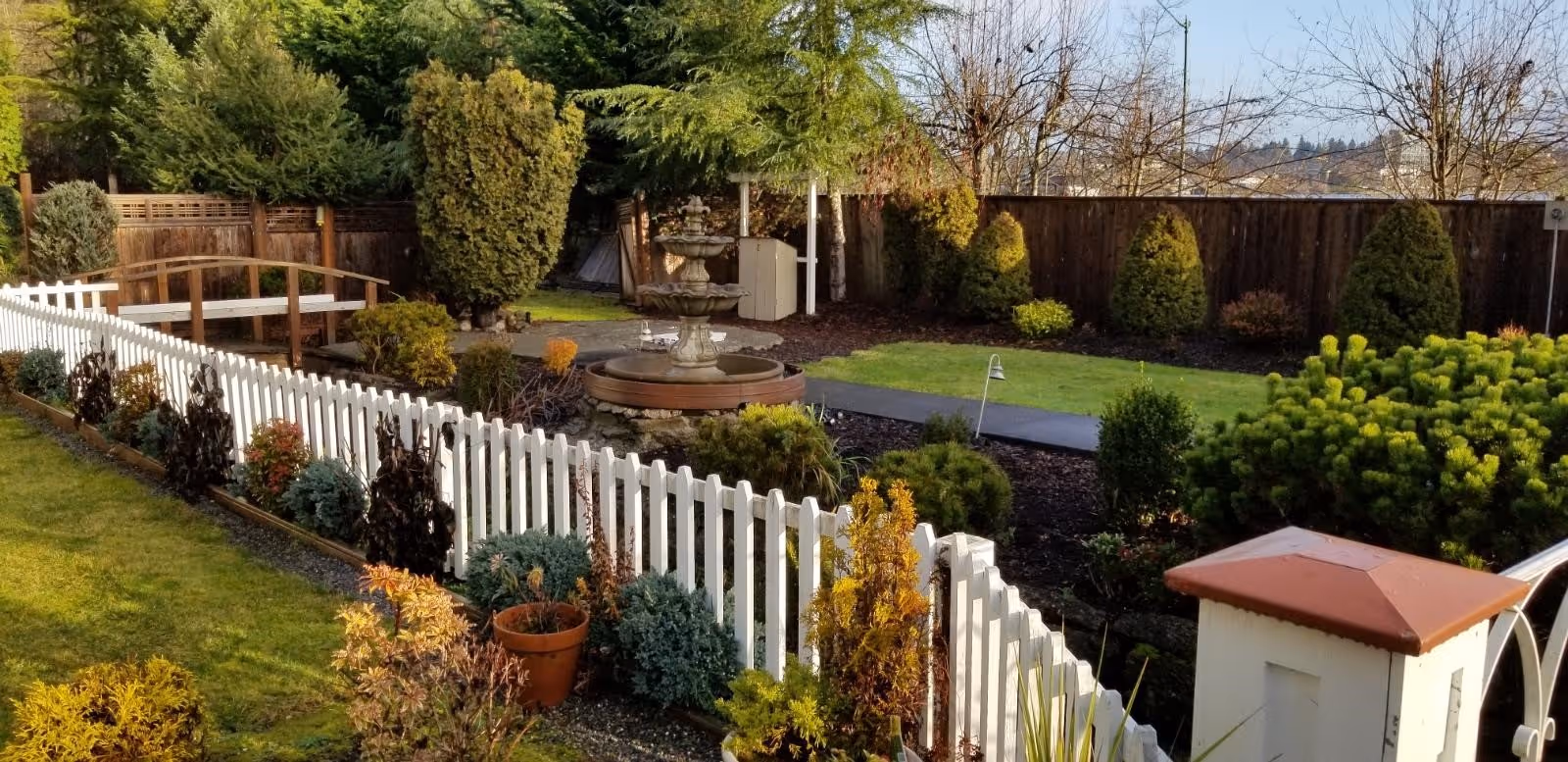 A well-maintained garden area with a white picket fence, various shrubs and plants, a small wooden bridge, and a multi-tiered water fountain surrounded by greenery and trees.