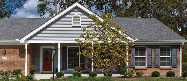 Single-story residential building with a gray shingled roof, gray siding, and brick accents. The front porch has white columns and a red door. There are several windows with dark shutters and a small tree and shrubs in the front yard.
