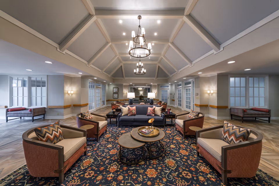 A spacious and elegantly decorated common area in a retirement community featuring a high vaulted ceiling with chandeliers, a central seating arrangement with a blue sofa and four armchairs around two round coffee tables on a patterned rug, and additional seating along the walls with large windows and soft lighting.