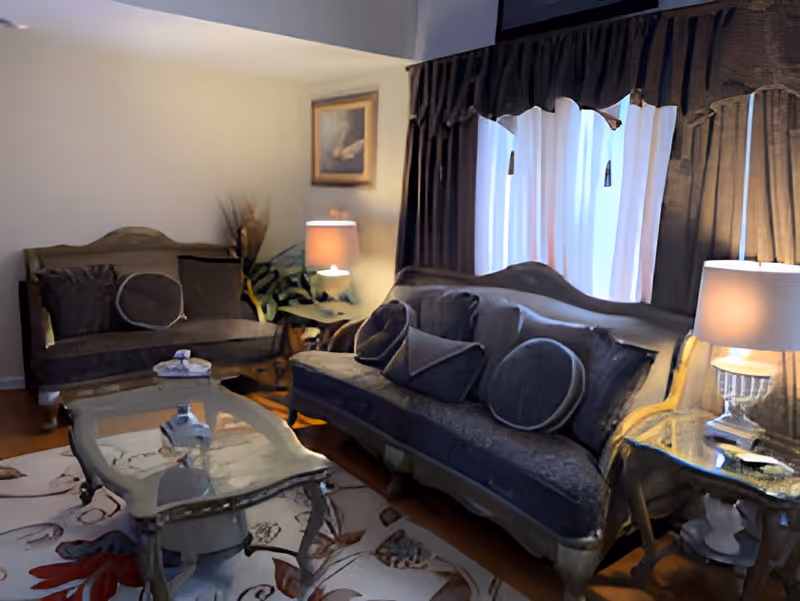 A cozy living room with two vintage-style sofas adorned with multiple cushions, a glass-top coffee table on a floral patterned rug, two side tables each with a lamp, a large window with dark curtains, and a framed picture on the wall.