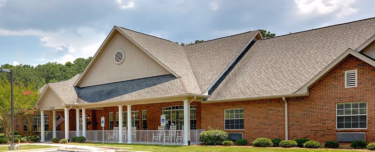 Exterior view of a single-story brick building with a large covered porch supported by white columns. The porch has several white rocking chairs and is surrounded by a white railing. There are multiple windows and a well-maintained lawn with shrubs in front. The sky is partly cloudy.