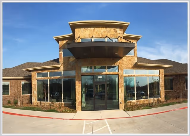 Front exterior view of a single-story building with large glass windows and a stone facade under a clear blue sky. The entrance has double glass doors with the number 5600 above them, and there is a covered awning above the entrance.