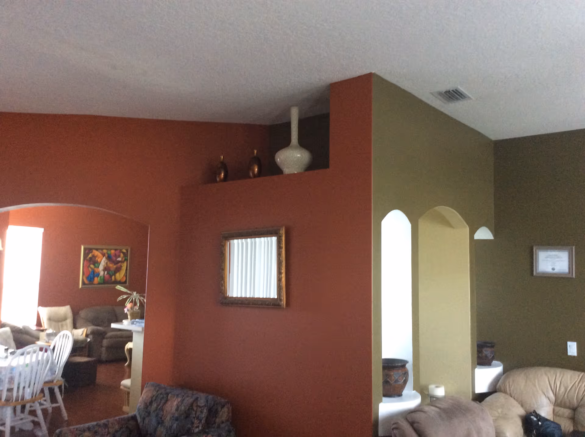 Interior view of a living area in a senior living facility with a mix of olive green and rust-colored walls. The room features a beige leather sofa, a floral patterned armchair, and a dining area with white chairs and a table covered with a tablecloth. Decorative vases are placed on a ledge above a framed mirror on the rust-colored wall. There is also a framed certificate on the olive green wall and some pottery displayed in wall niches.