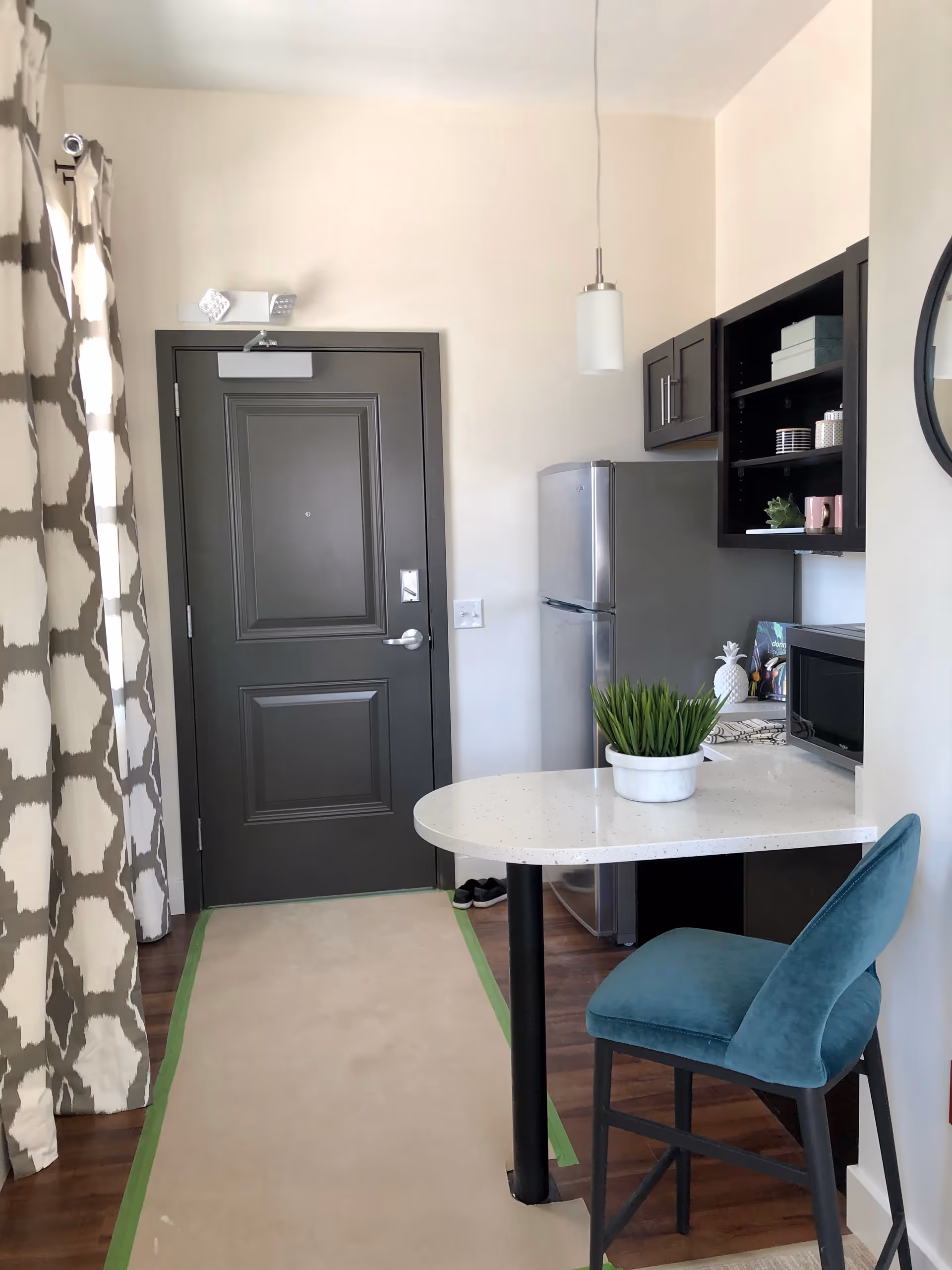 Interior view of a small kitchen area with a dark gray door, a stainless steel refrigerator, dark wood cabinets, a white countertop with a potted plant, a teal upholstered chair, and patterned curtains on the left side.