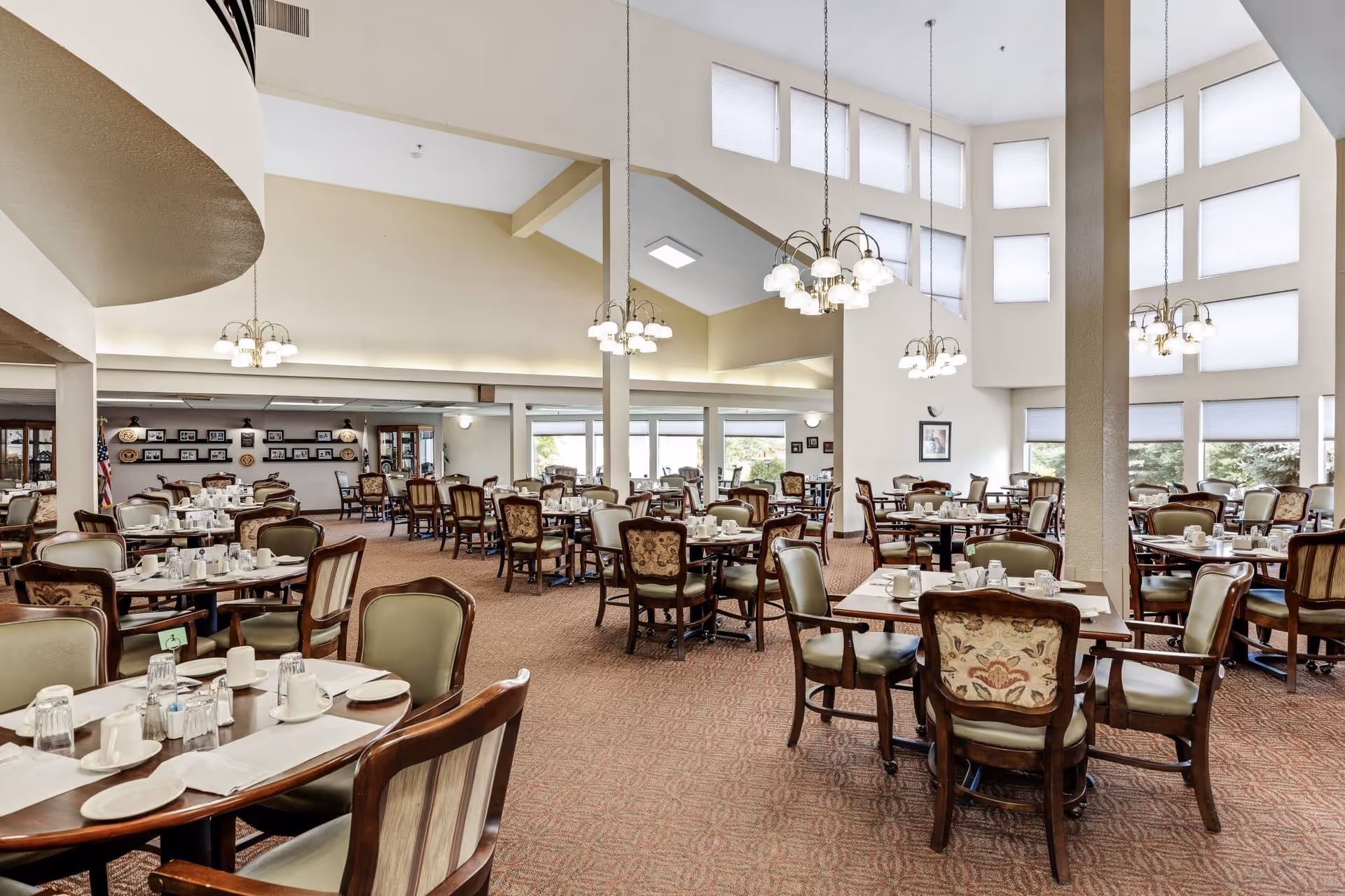 Spacious dining room with multiple tables and chairs arranged neatly. Each table is set with white cups, plates, glasses, and napkins. The room features high ceilings with large windows covered by blinds, allowing natural light to fill the space. Elegant chandeliers hang from the ceiling, and the carpet is patterned in warm tones. The walls are decorated with framed pictures and shelves displaying decorative items.