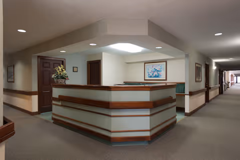 Interior view of a senior living facility reception area with a wooden and frosted glass front desk. The hallway extends to the right with doors and framed artwork on the walls. The ceiling has recessed lighting and a skylight above the desk.