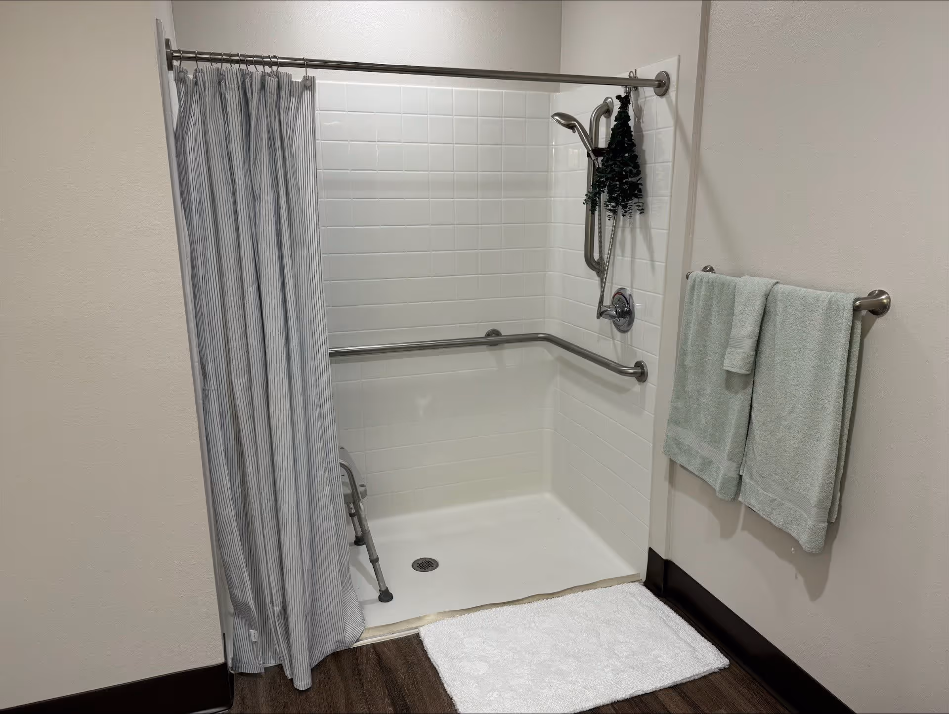 A walk-in shower with white tiled walls and a silver grab bar along two sides. There is a silver adjustable showerhead and a small hanging plant. A striped shower curtain is partially drawn to the left. A white bath mat is placed on the dark wood floor outside the shower. Two light green towels hang on a silver towel rack mounted on the wall to the right.