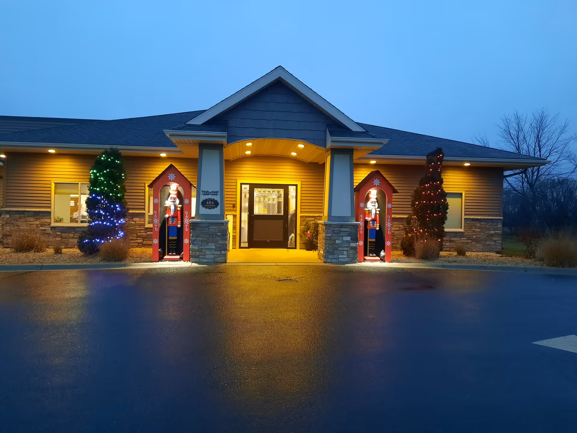 Exterior view of a single-story building at dusk with warm yellow lights illuminating the entrance. Two large decorative nutcracker figures stand in red shelters on either side of the doorway. Small trees decorated with colorful lights flank the entrance. The building has beige siding with stone accents and a dark roof.