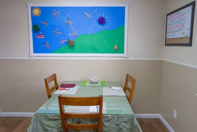 A small dining area with a square table covered by a green tablecloth and four wooden chairs. The table is set with placemats, napkins, and utensils. On the wall above the table is a colorful bulletin board decorated with a sun, dragonflies, and pom-poms. To the right, there is a framed whiteboard with writing on it.