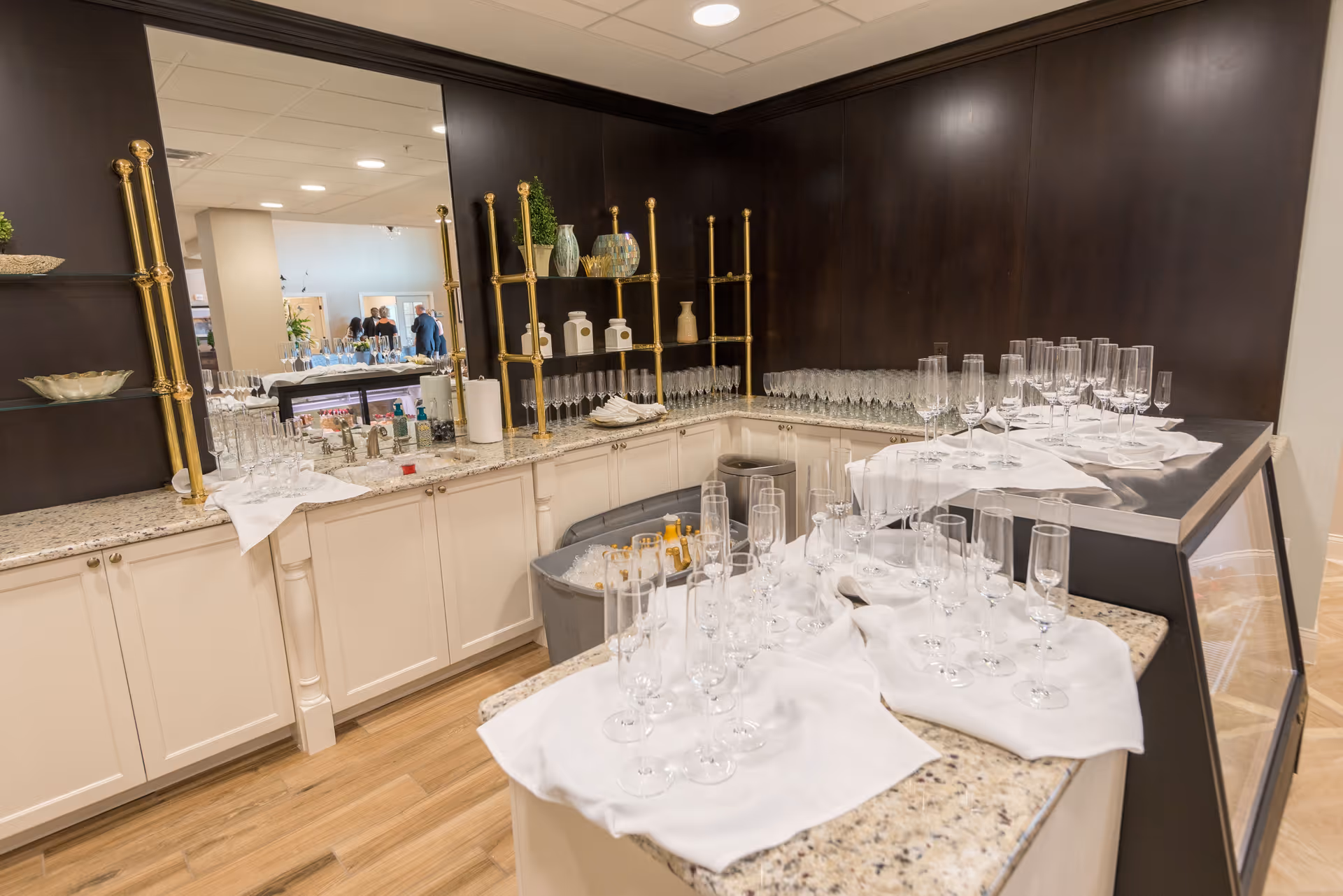 A countertop area with numerous empty champagne flutes arranged on white cloths. The space features light-colored cabinets, a granite countertop, and a large mirror with gold-colored decorative shelving. In the background, a few people are visible in a well-lit room.