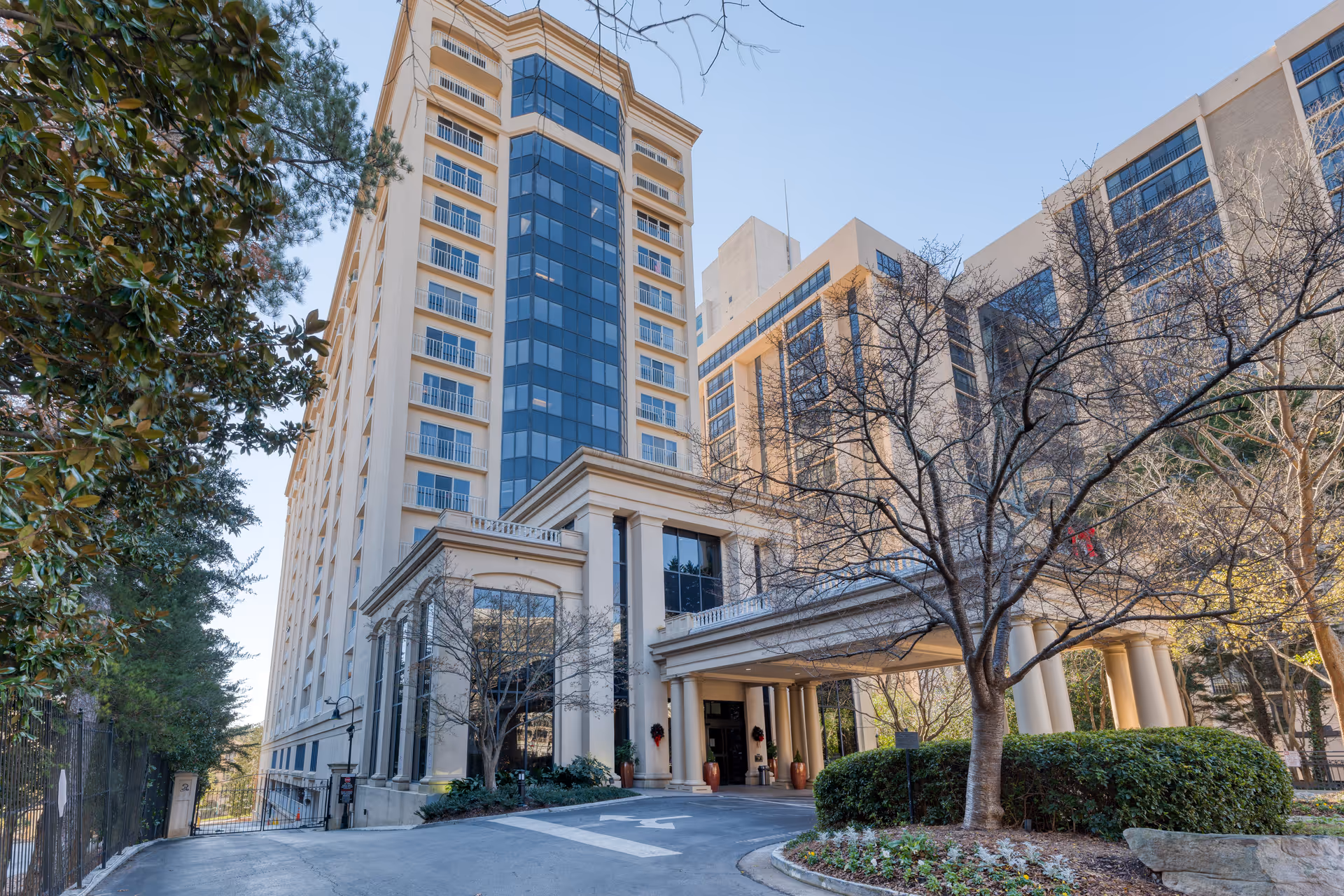 Exterior view of a tall, multi-story building with large windows and balconies, surrounded by trees and landscaping. The entrance features a covered driveway with columns and potted plants. The sky is clear and blue.