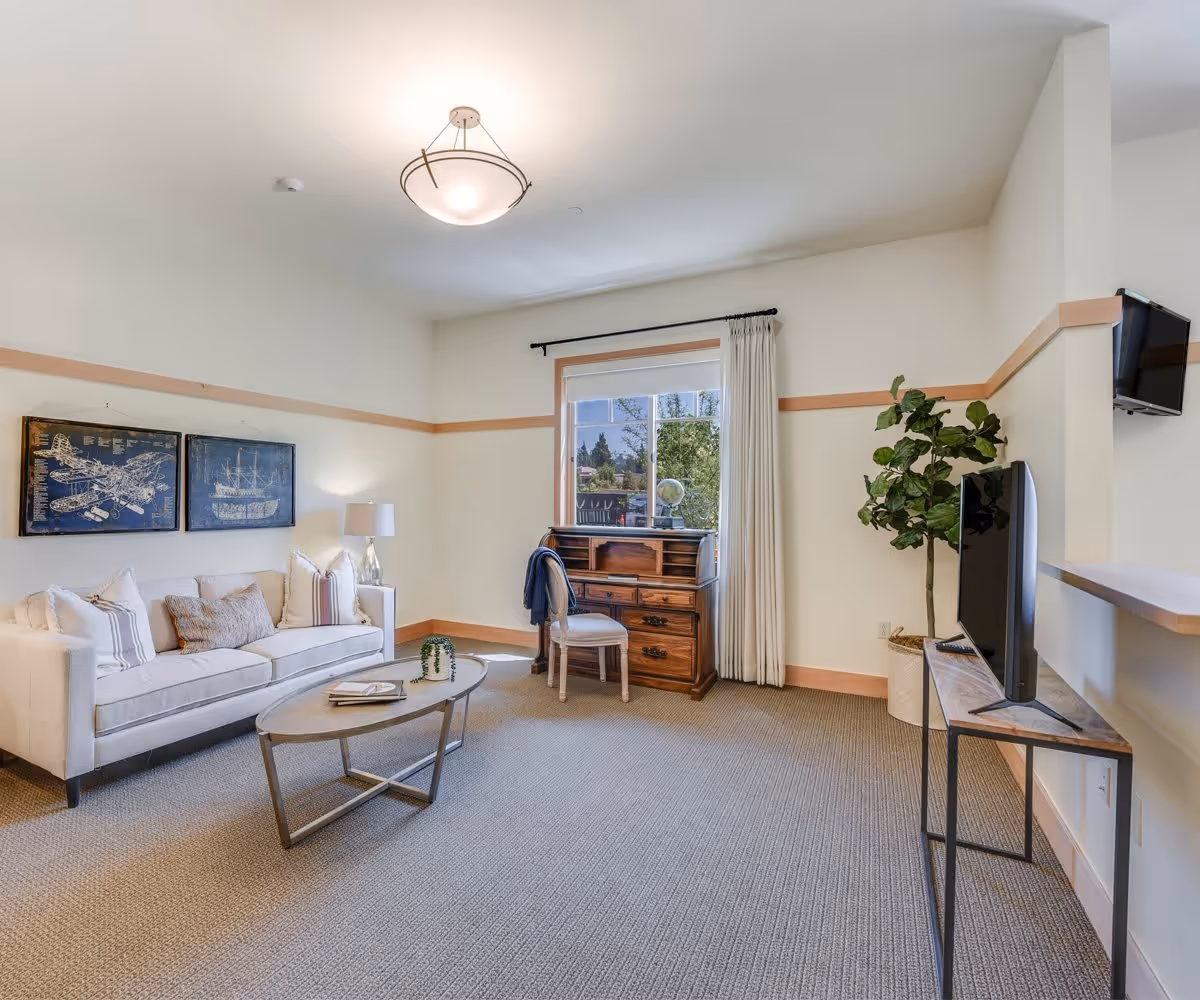 A bright and spacious living room with a beige sofa adorned with pillows, a wooden coffee table with books and a small plant, a wooden desk with a chair near a window with white curtains, a flat-screen TV on a stand, a potted plant, and two framed artworks on the wall.