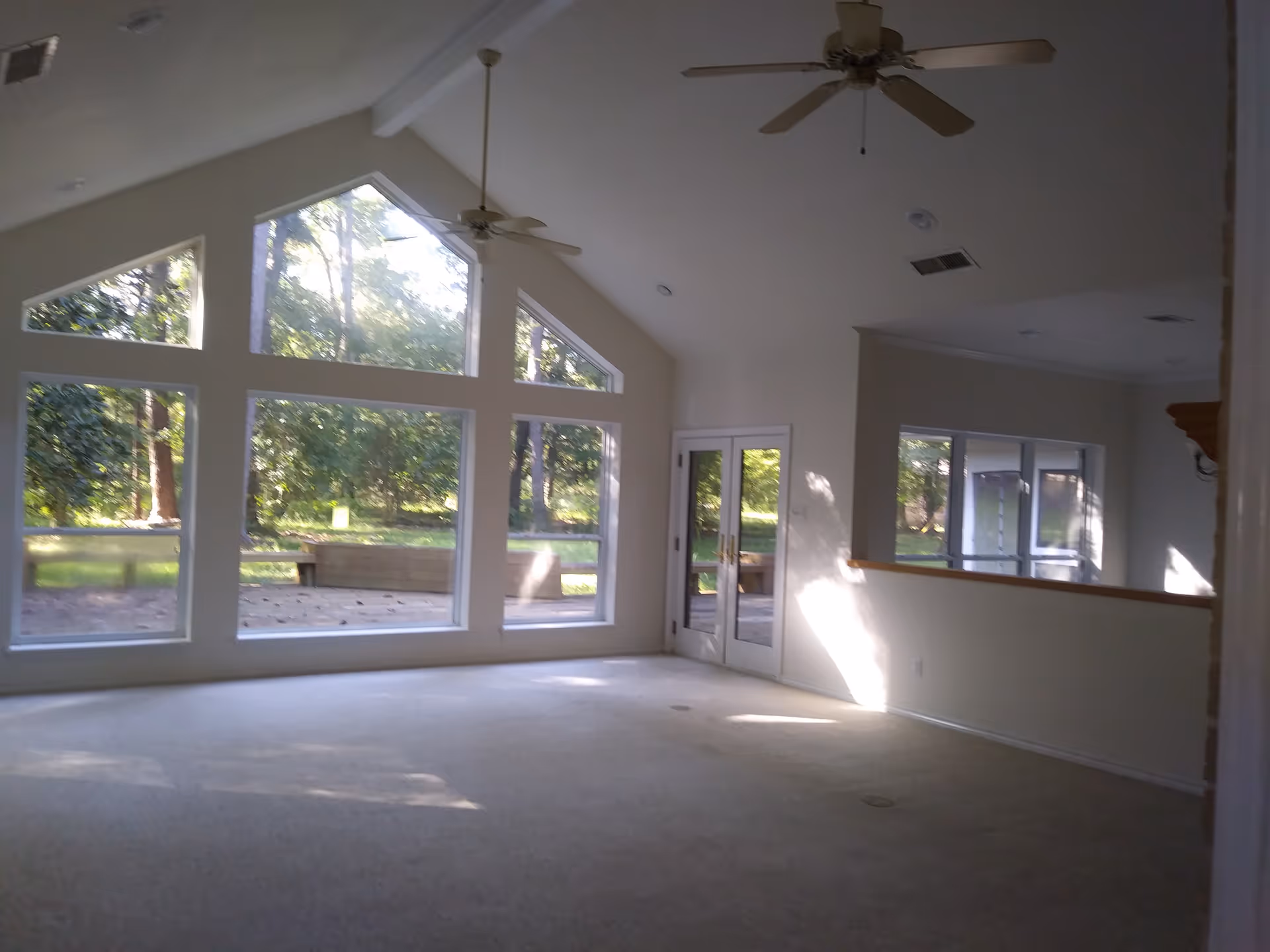 Spacious empty living room with vaulted ceiling, large multi-pane windows and ceiling fans overlooking a wooded yard.