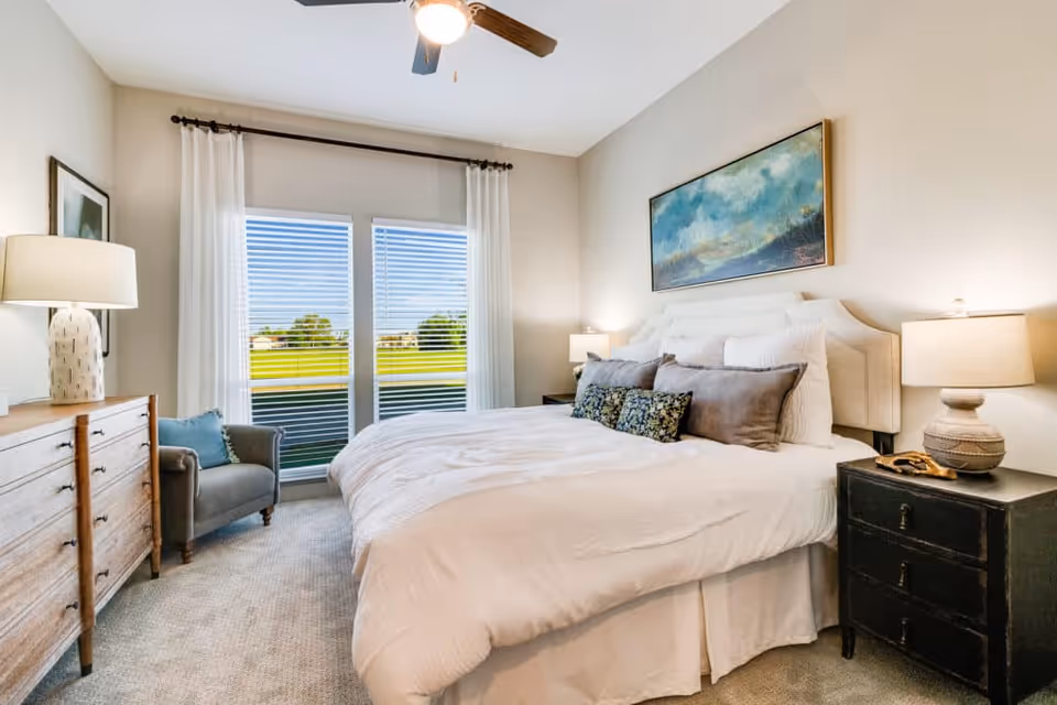 Bright, neatly staged bedroom with a large upholstered bed, nightstands, dresser, armchair, and windows overlooking a green lawn.