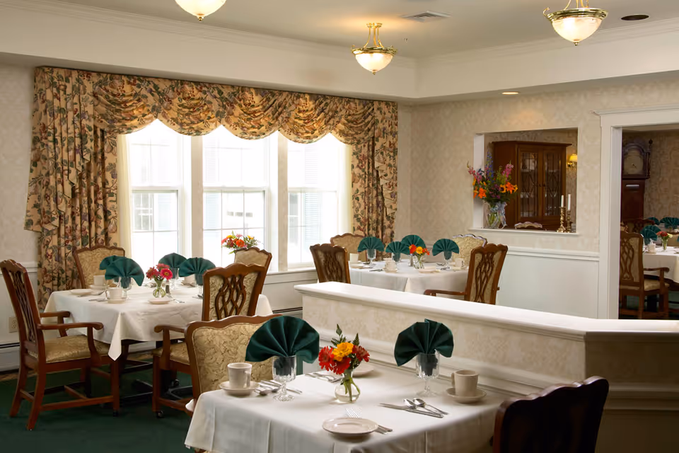 A dining room with several tables covered in white tablecloths, each set with cups, plates, silverware, and green folded napkins. Floral arrangements are placed in small vases on each table. The room has large windows with floral-patterned curtains and classic wooden chairs with upholstered seats. The walls are decorated with light wallpaper and there are ceiling lights providing warm illumination.
