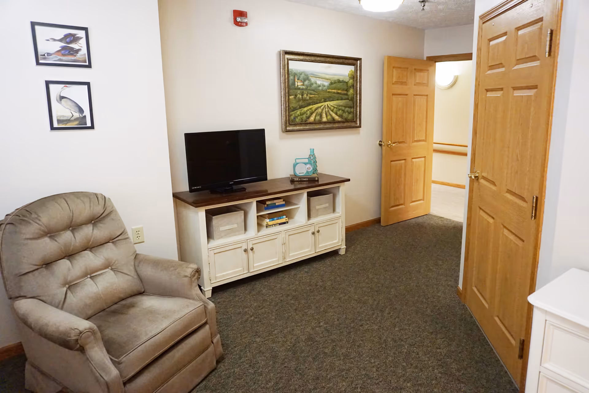 A cozy living room area in Snyder Village Assisted Living featuring a beige recliner chair, a white TV stand with a flat-screen TV, books, and decorative items. The walls are adorned with framed artwork of birds and a landscape painting. Two wooden doors are visible, one open leading to a hallway.
