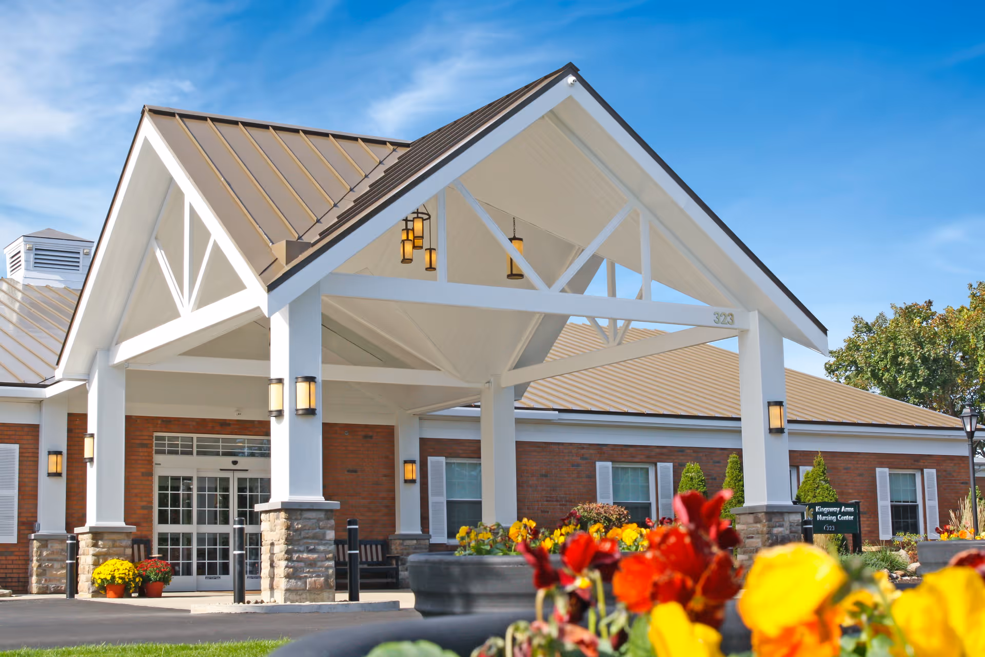 Front entrance of a brick nursing center with a covered porte-cochere, exterior lights, and colorful flowers in the foreground.
