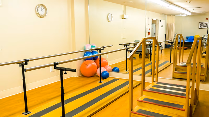 A physical therapy or rehabilitation room with parallel bars for walking exercises, a set of stairs with handrails, several colorful exercise balls, a large wall mirror, and a clock on the wall.