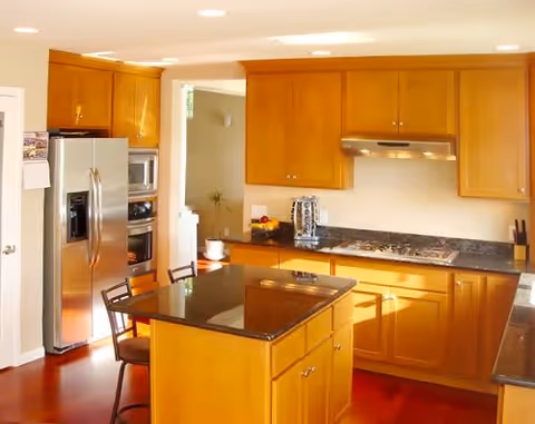 A bright kitchen with wooden cabinets, a stainless steel refrigerator, built-in microwave and oven, a black countertop island with two chairs, and a gas stove with a range hood.