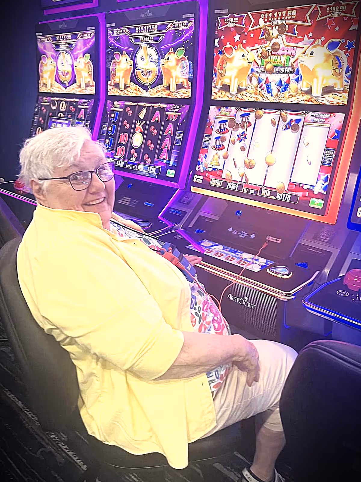 An elderly woman with white hair and glasses, wearing a yellow jacket and floral shirt, is sitting and smiling while playing on brightly lit slot machines in a gaming area.