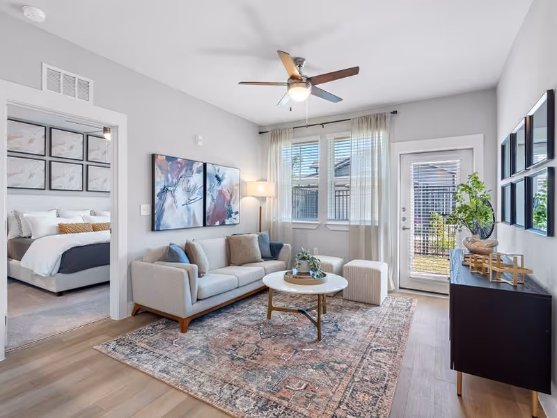 Bright contemporary living room with a sofa, round coffee table on a patterned rug, wall art, ceiling fan, and a view into a bedroom and a glass door to the outside.