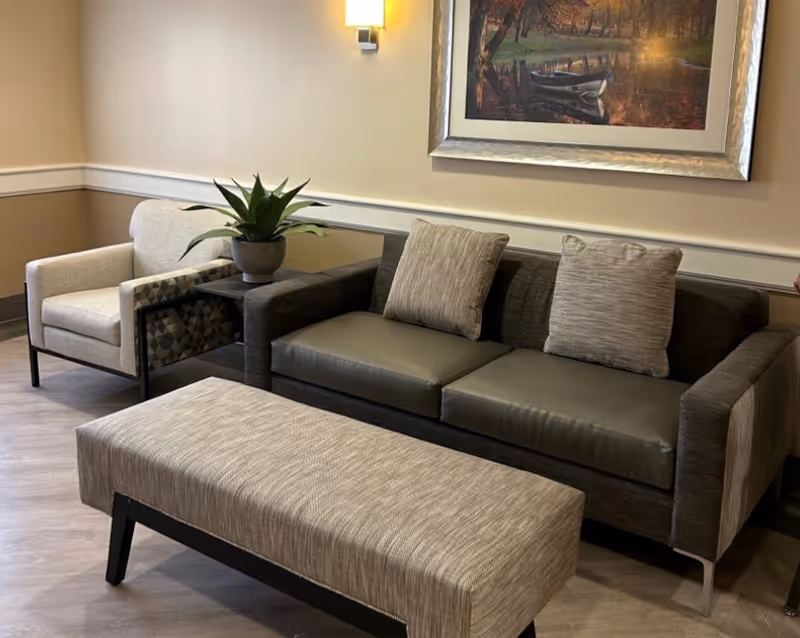 A cozy seating area with a dark gray sofa featuring two light gray cushions, a matching armchair with patterned sides, a rectangular upholstered ottoman, and a small side table with a potted plant. A framed picture of a boat on a lake hangs on the beige wall above the sofa, illuminated by a wall-mounted light fixture.