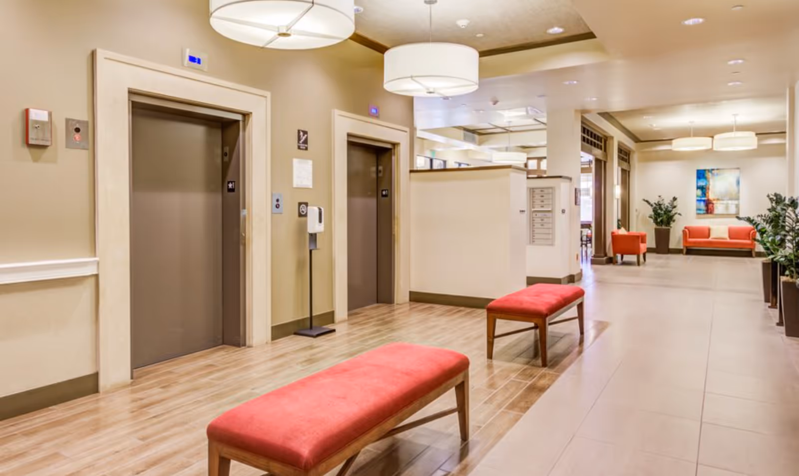 Bright lobby interior with two elevators, red upholstered benches, seating area, plants, and pendant lights.