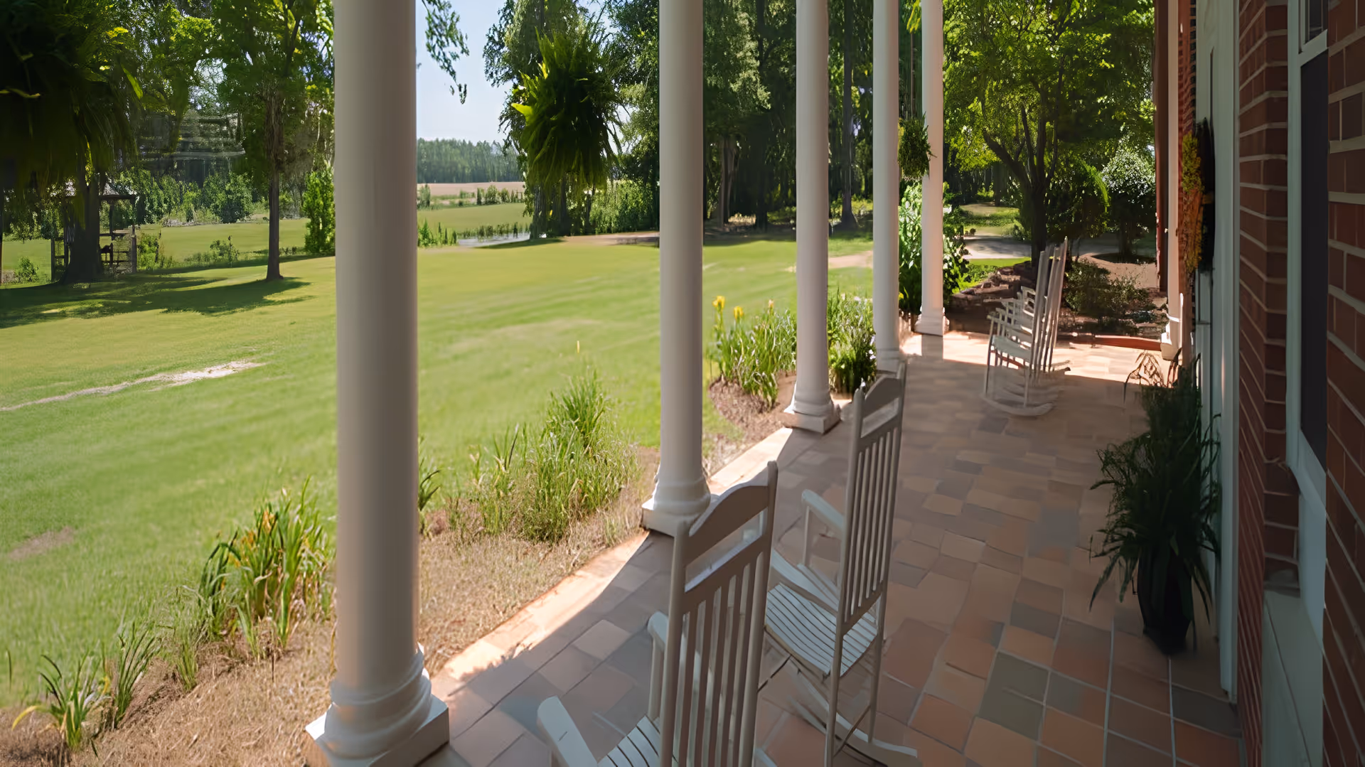 A covered porch with white rocking chairs overlooking a green lawn and trees. The porch has tiled flooring and white columns supporting the roof. There are plants along the edge of the porch and a brick wall on one side.