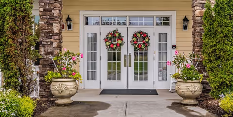 Entrance to a building with double white doors decorated with two floral wreaths. The doorway is flanked by stone pillars and large planters with pink flowers. There are two black wall-mounted lanterns on either side of the doors and greenery surrounding the entrance.