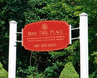 A red sign with white posts reading 'Rose Tree Place An Independent and Assisted Living Residence Wellness Program for the Memory Impaired 610-565-1405' surrounded by green trees and bushes.